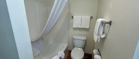 Et lille, rent badeværelse med et hvidt toilet, et hvidt badekar og en bruseforhæng. Der hænger to hvide håndklæder på væggen.
