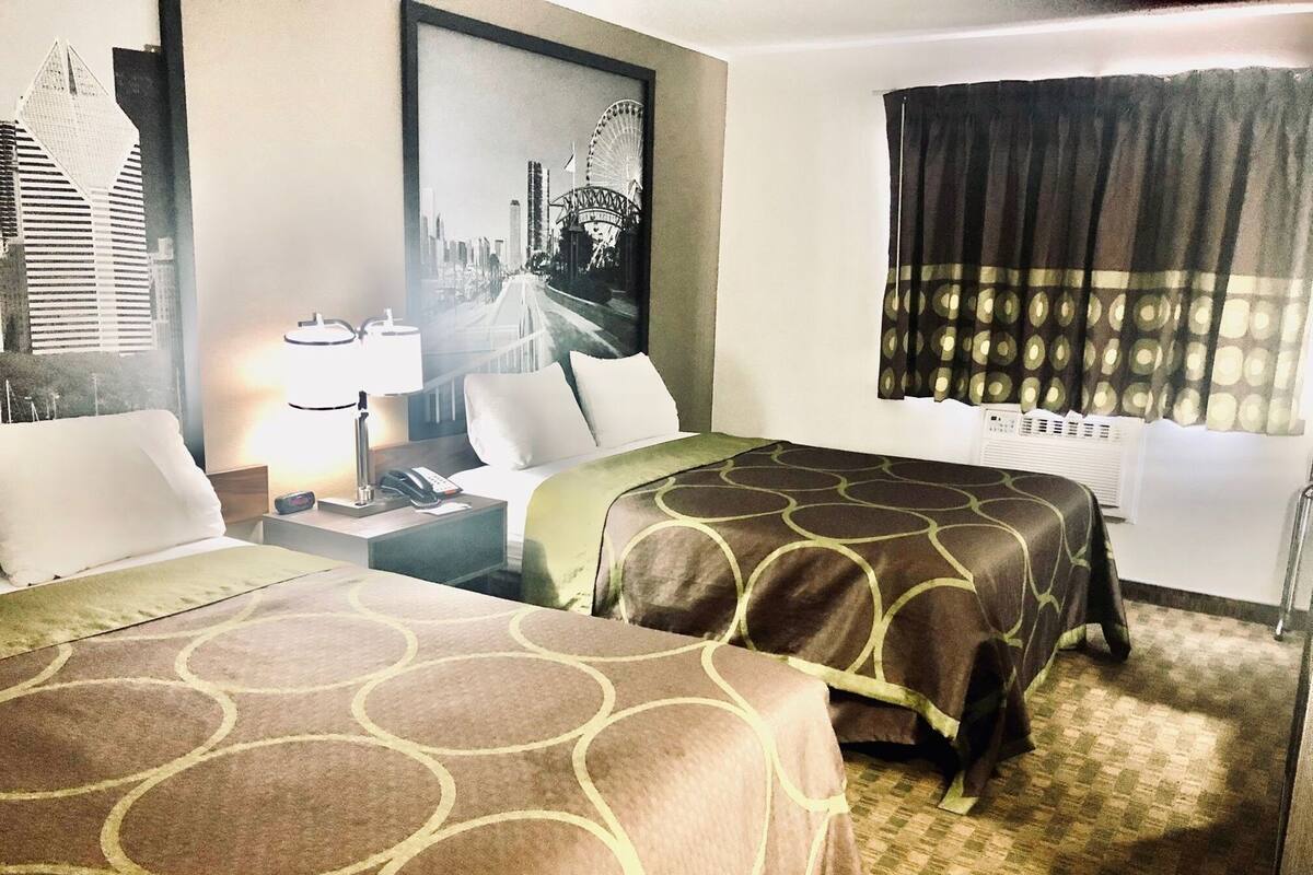 Habitació d'hotel amb dos llits, una tauleta de nit, una làmpada, un quadre amb una vista de la ciutat i una finestra amb cortines.