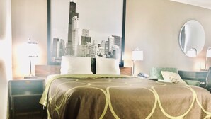 Habitación de hotel con una cama grande, dos mesitas de noche, vistas a la ciudad, un espejo redondo y una silla verde.