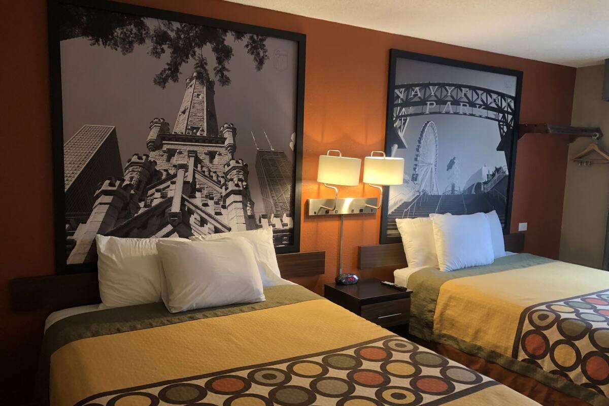 Una habitació d'hotel amb dos llits, un quadre gran de paisatge urbà a la paret, una tauleta de nit amb una làmpada i un penjallabecs.