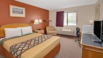 A hotel room with a bed, a desk with a computer, a chair, a window with curtains, and a wall-mounted air conditioner.