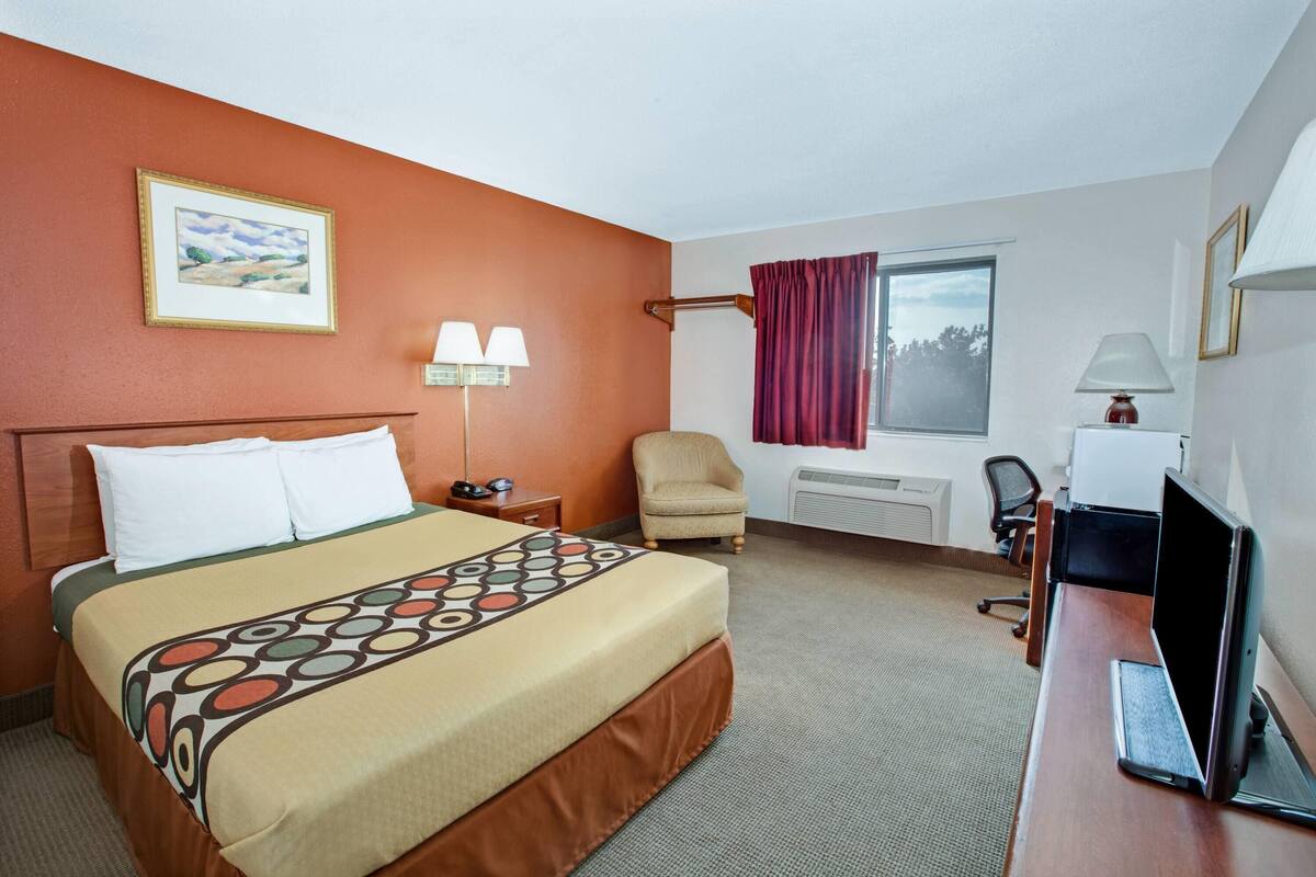 Una habitació d'hotel amb un llit, un escriptori amb un ordinador, una cadira, una finestra amb cortines i una làmpada penjada a la paret.