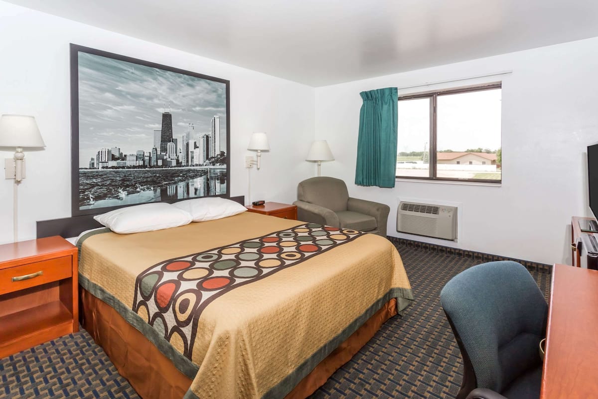 Hotelkamer met een bed, een bureau, een stoel, een raam met gordijnen, een wandairconditioner en een stadsgezichtschilderij.