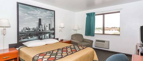 Habitación de hotel con cama, escritorio, silla, ventana con cortinas, aire acondicionado montado en la pared y una pintura de un paisaje urbano.