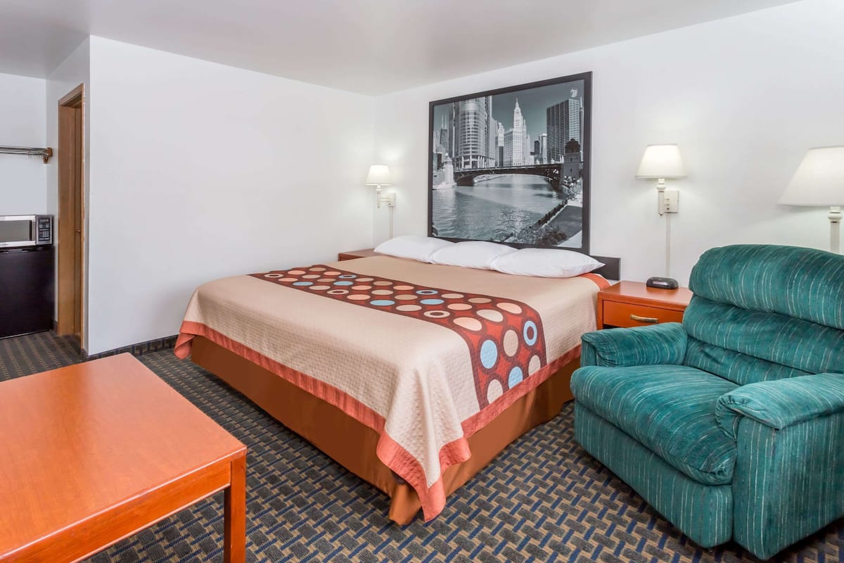 Hotelkamer met een bed, een groene fauteuil, een houten tafel, een aan de muur bevestigde televisie en een stadsgezicht schilderij.