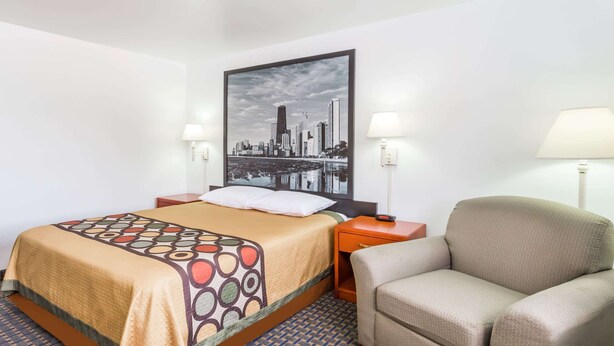 Een hotelkamer met een bed, een stoel, een nachtkastje, een lamp en een stadslandschap schilderij aan de muur.