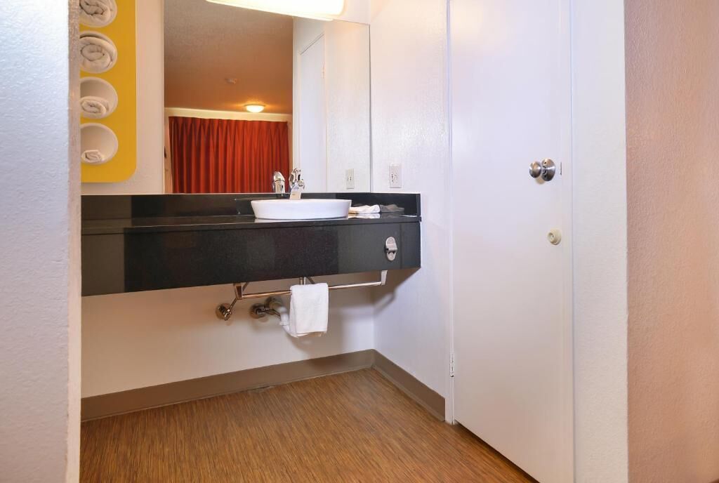 A bathroom with a black countertop, white sink, and a mirror. There is a towel rack with rolled towels and a red curtain in the background.