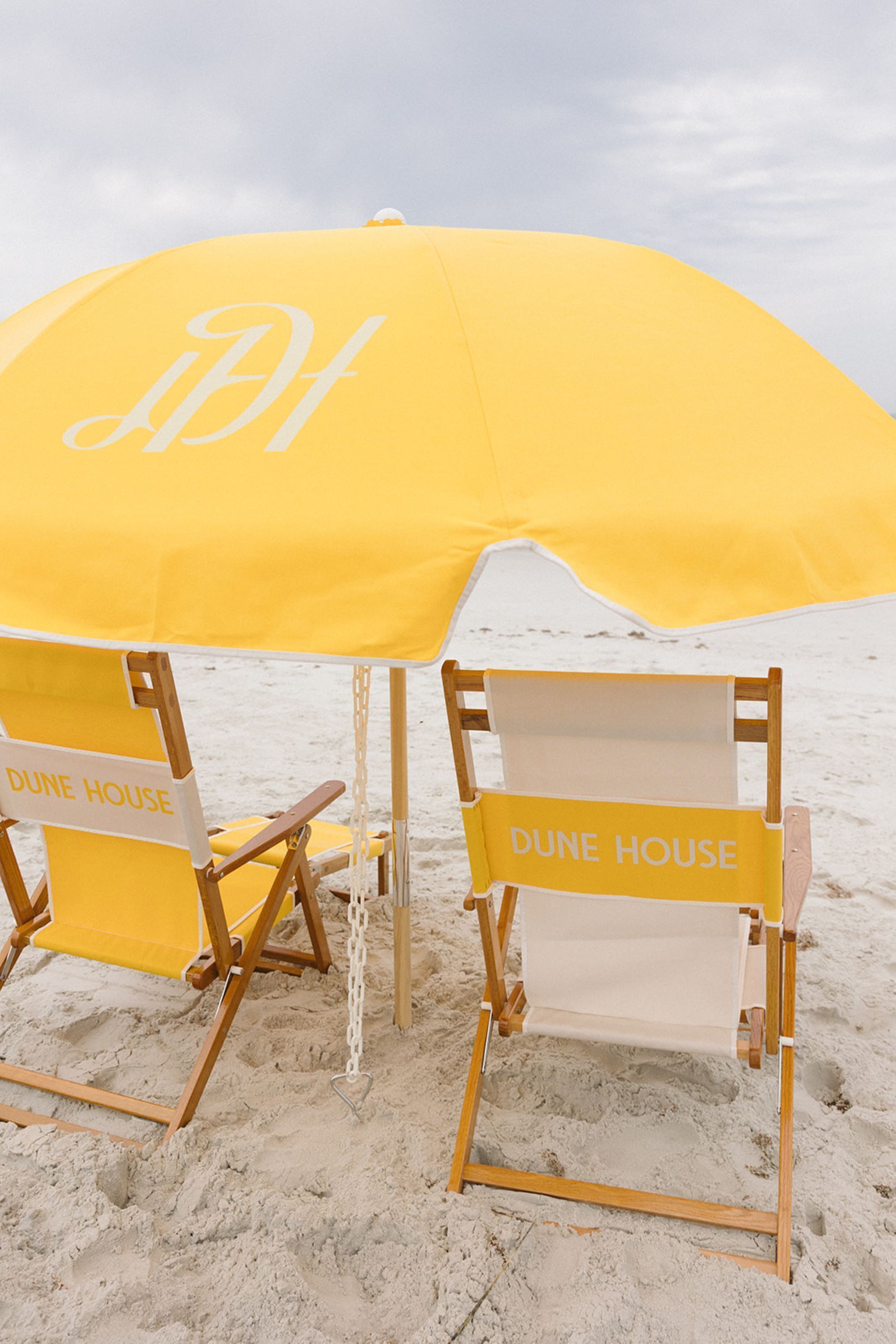 Two beach chairs with 'DUNE HOUSE' labels, under a yellow umbrella on a sandy beach.