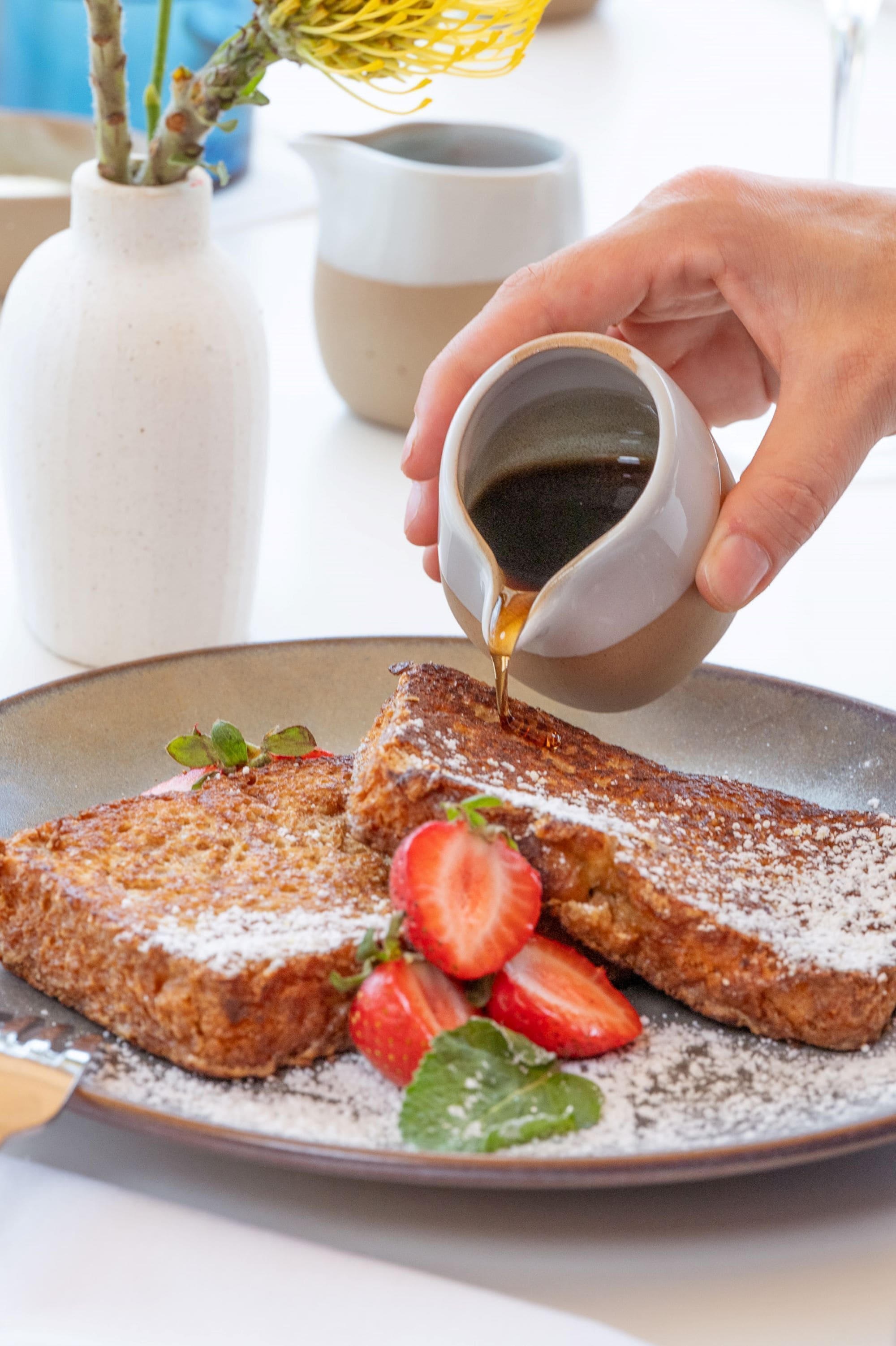 A hand pouring syrup over French toast with strawberries on a plate.