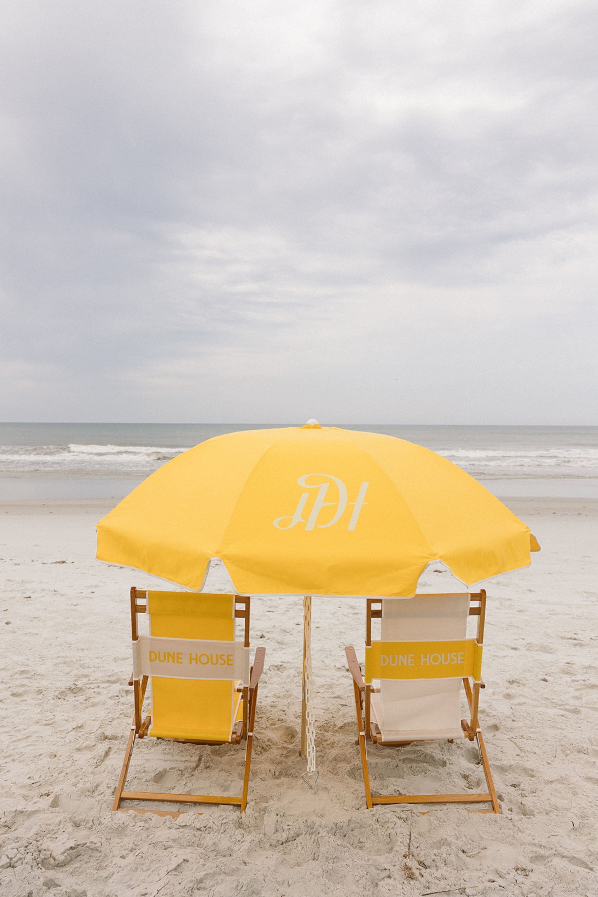 Two beach chairs with 'DUNE HOUSE' signage, under a yellow umbrella on a sandy beach.