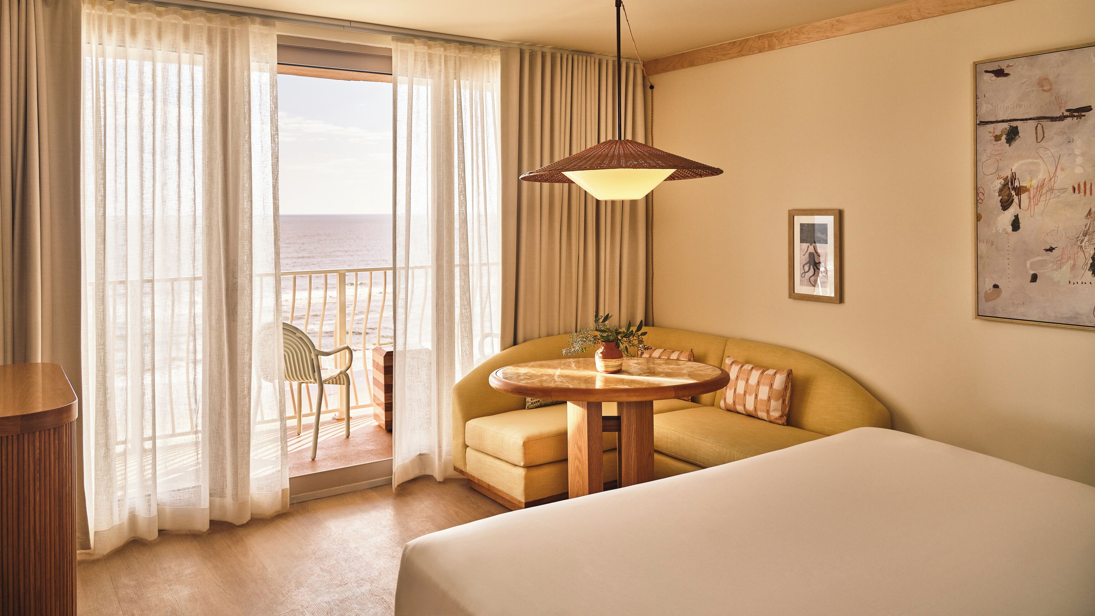 Une chambre d’hôtel avec un lit, une table ronde en bois, un canapé, une chaise, un balcon avec des rideaux et un tableau au mur.