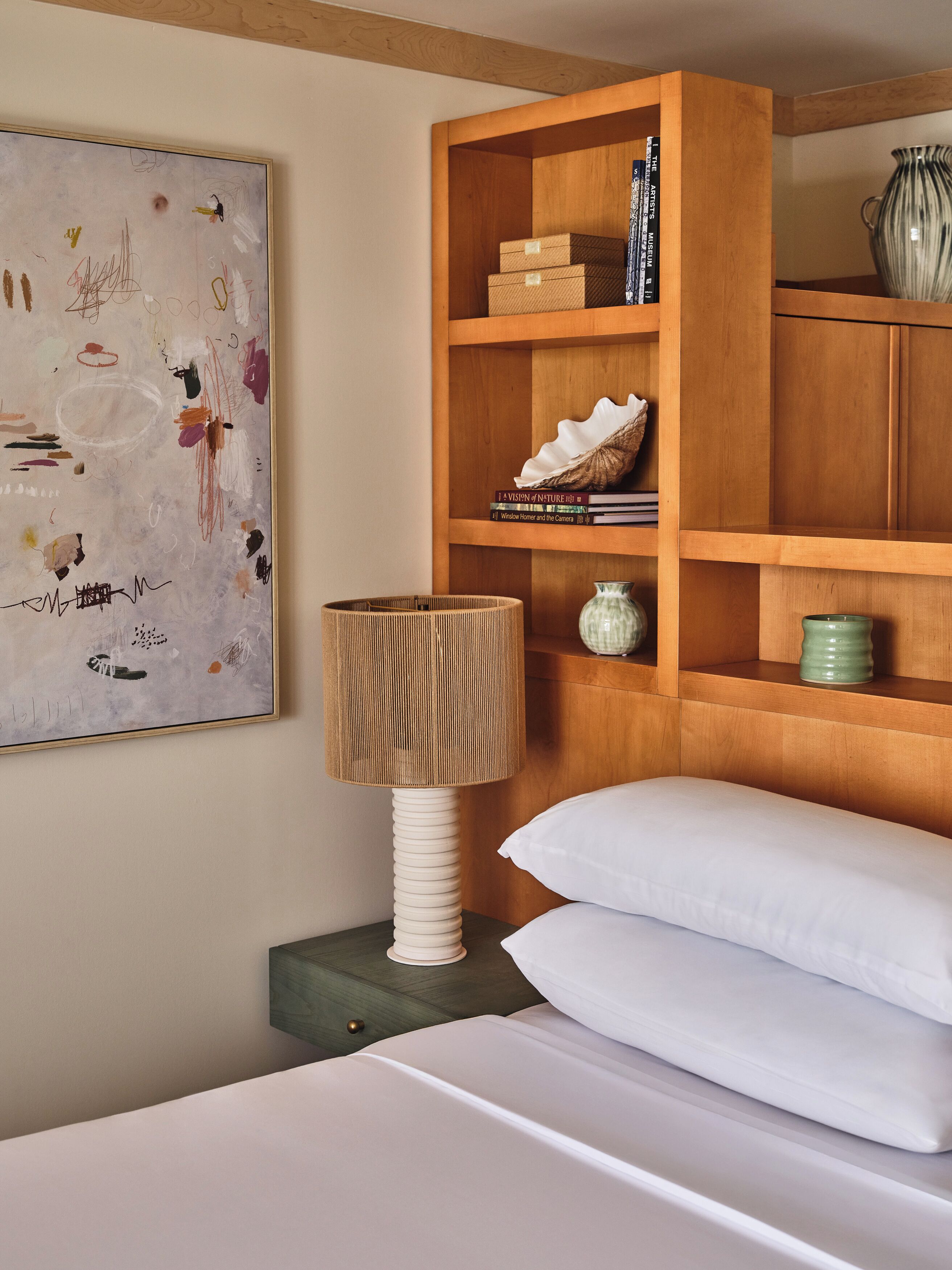 A neatly arranged bedroom with a wooden headboard, a bedside lamp, a white bed, and a shelf with books and decorative items.