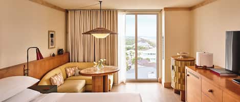 A hotel room with a bed, a sofa, a dining table, a TV, and a view of the city.