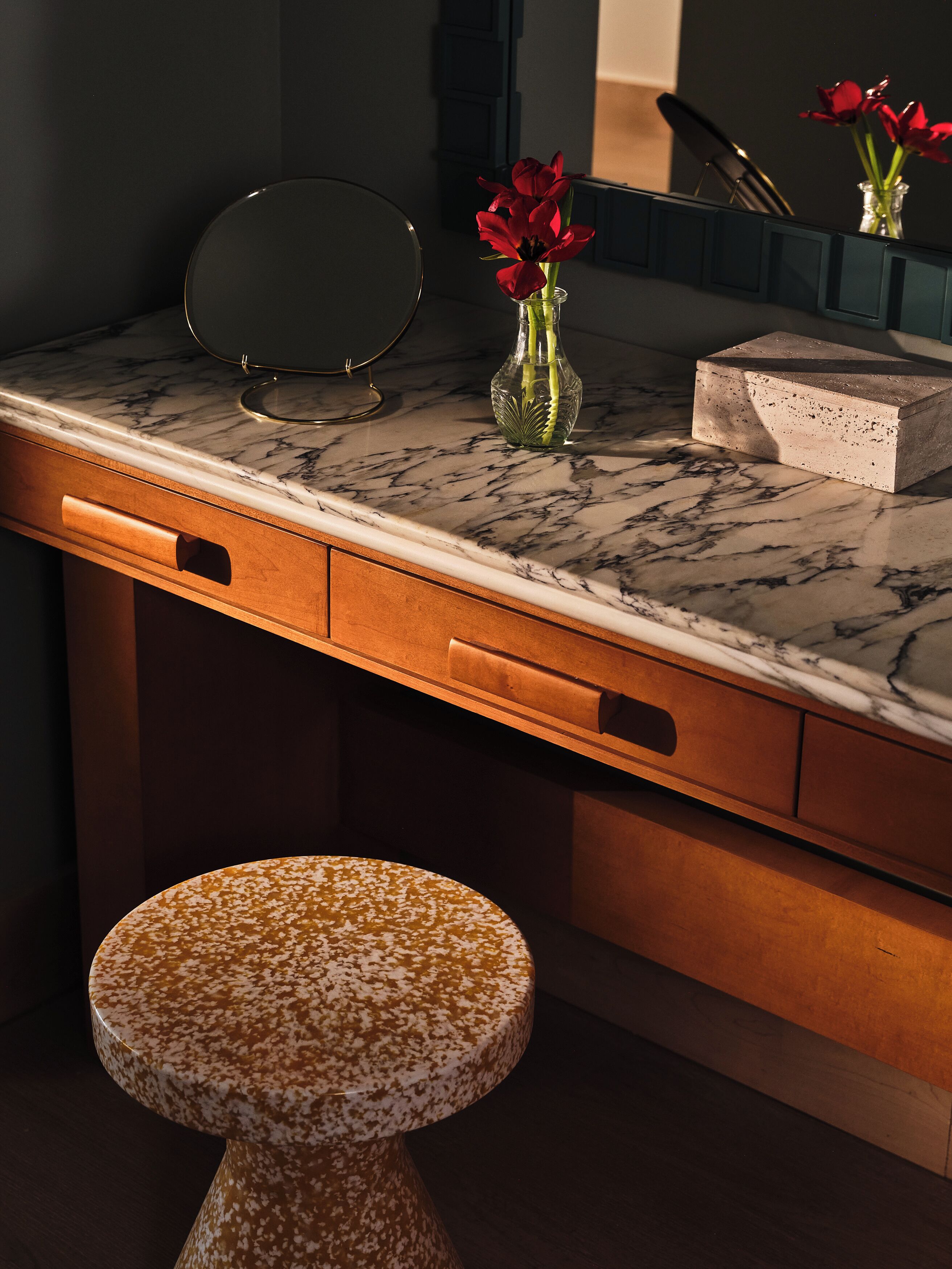 A bathroom vanity with a marble countertop, a glass vase with red flowers, and a round stool.
