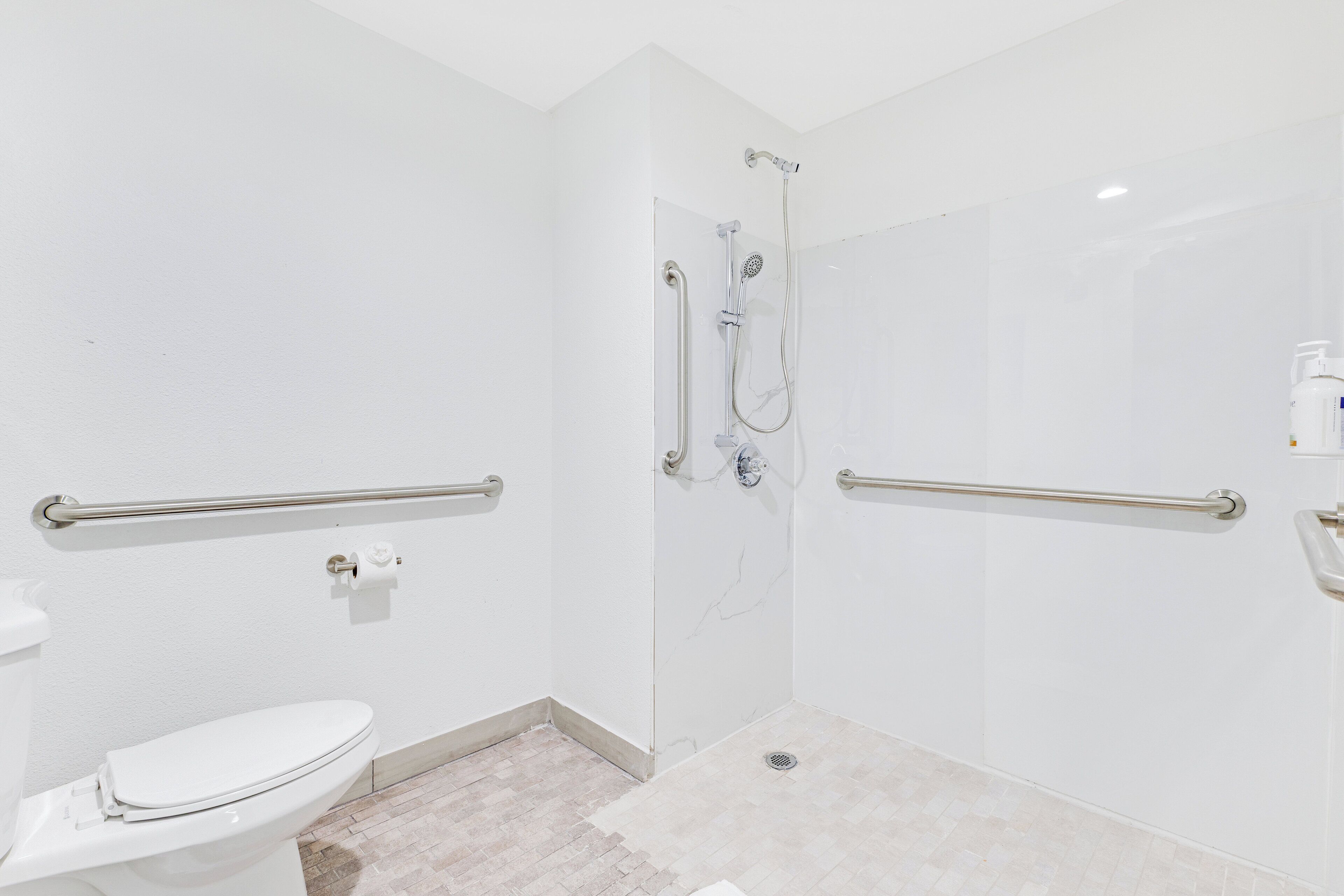 A bathroom with a toilet, a shower with grab bars, and a white tiled floor.