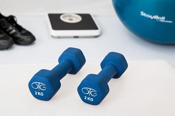Dos mancuernas azules con la etiqueta 2KG sobre una superficie blanca, una balanza digital y una pelota de ejercicios azul al fondo.