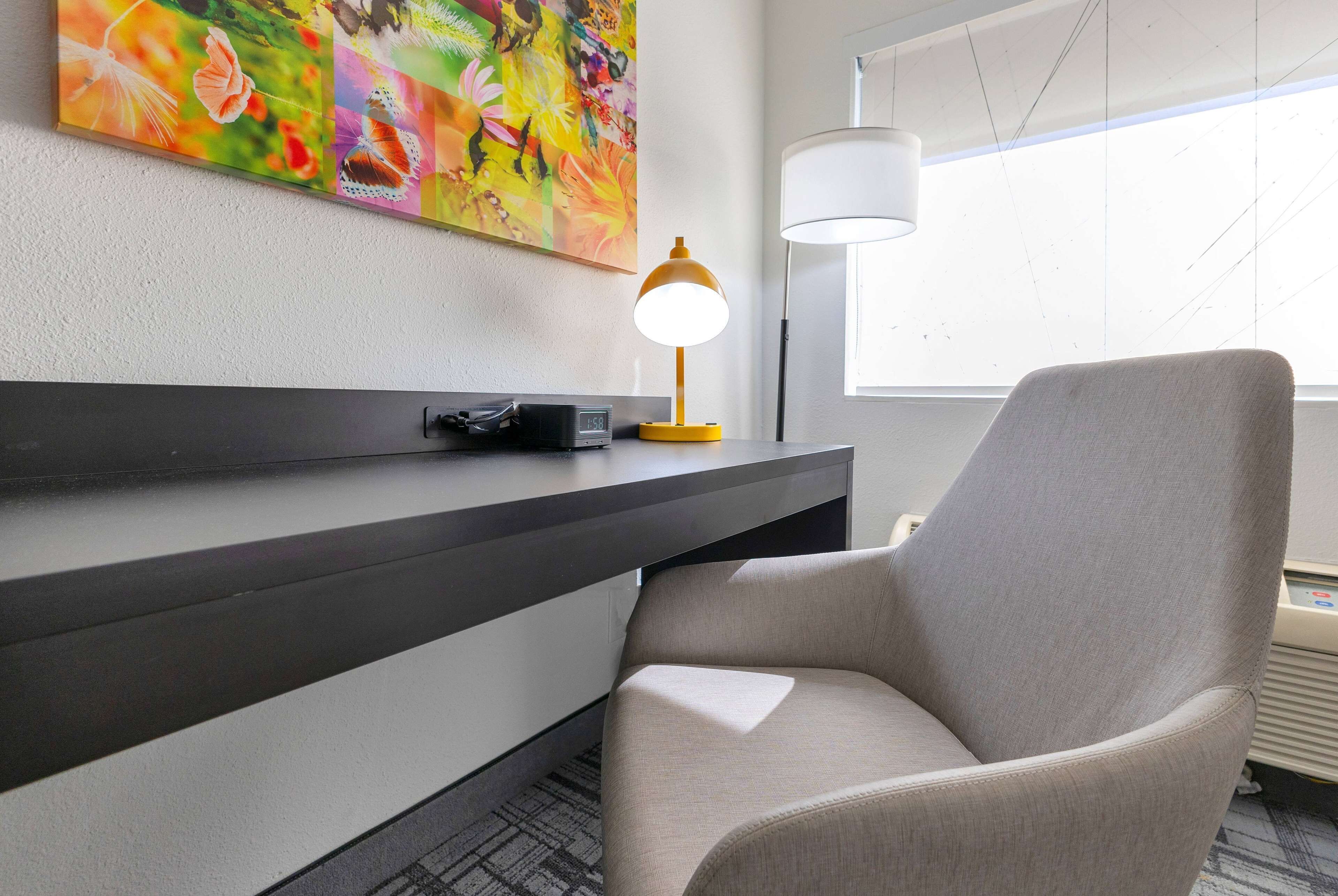 A modern hotel room with a grey armchair, a desk with a lamp, and a colorful abstract painting on the wall.