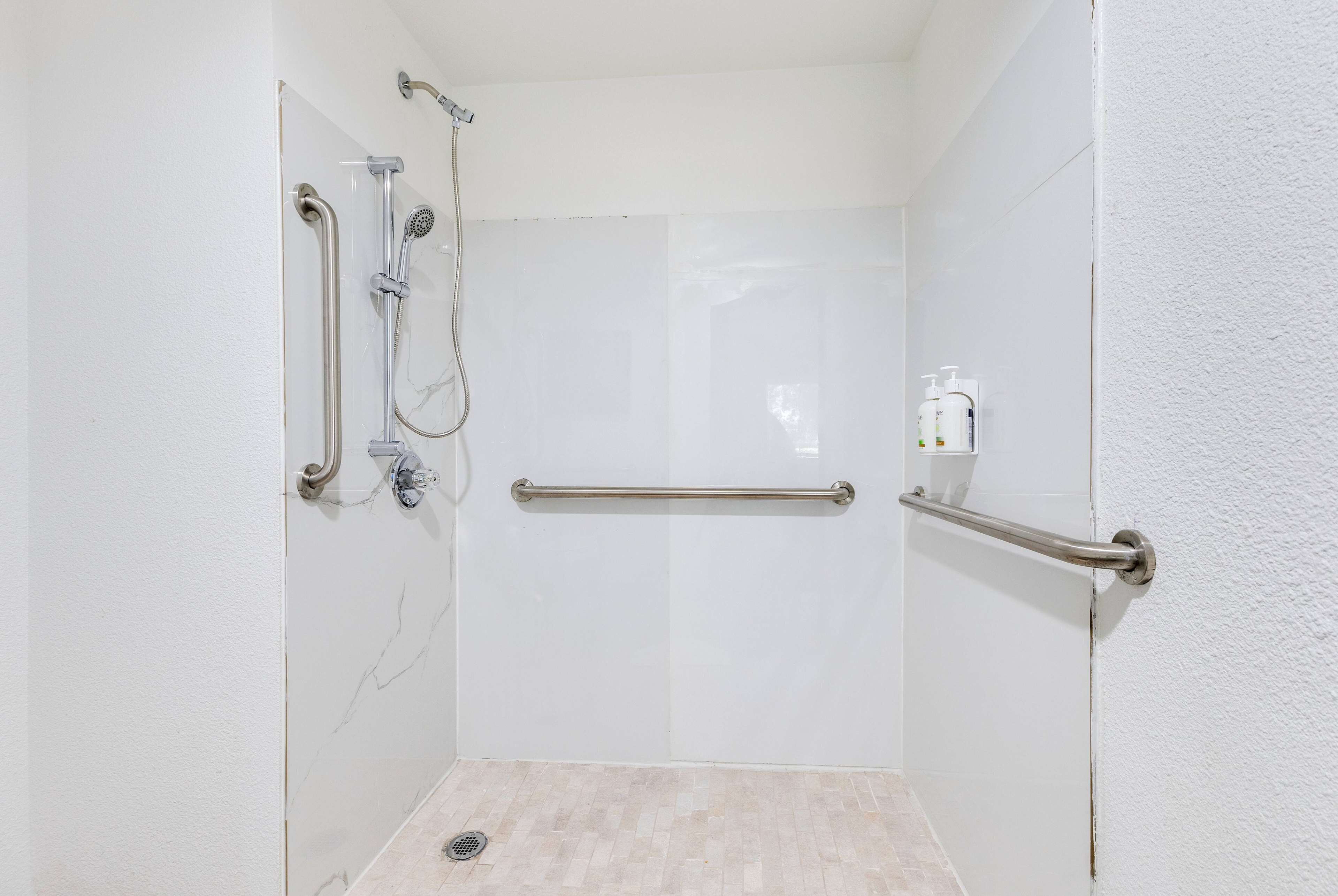 Casa de banho pequena e branca com azulejos, chuveiro, barras de apoio e secador de mãos.