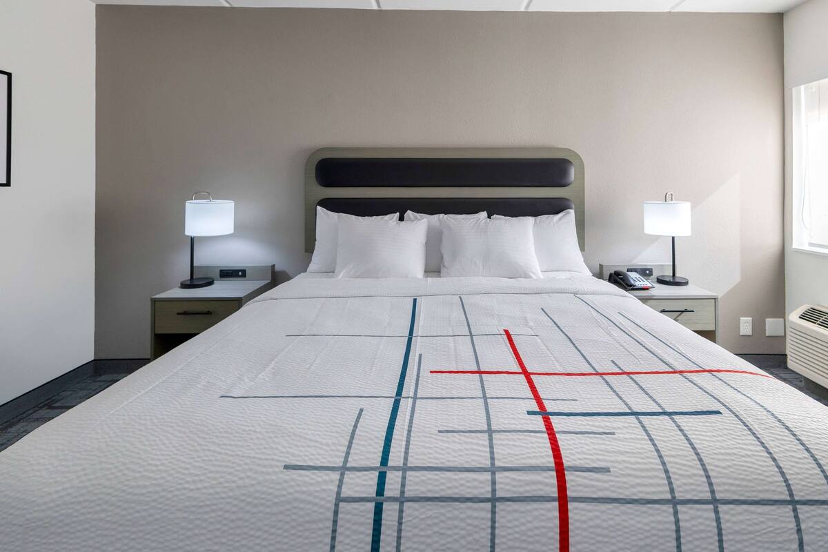 A hotel room with a large bed, two bedside tables with lamps, a wall-mounted heater, and a window with curtains.