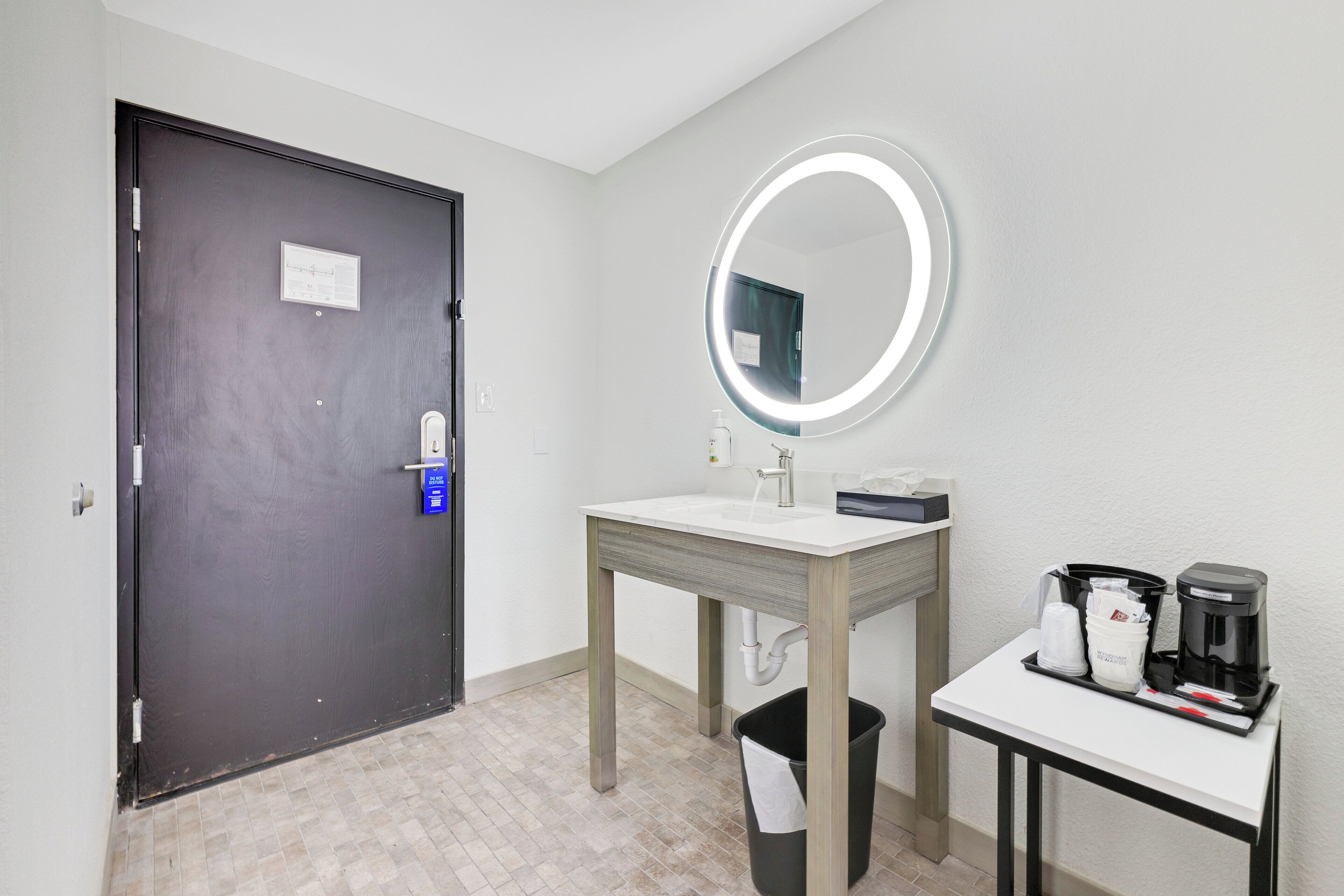 A hotel room with a dark door, a round mirror with a light, a sink, a small table with a coffee maker, and a trash bin.