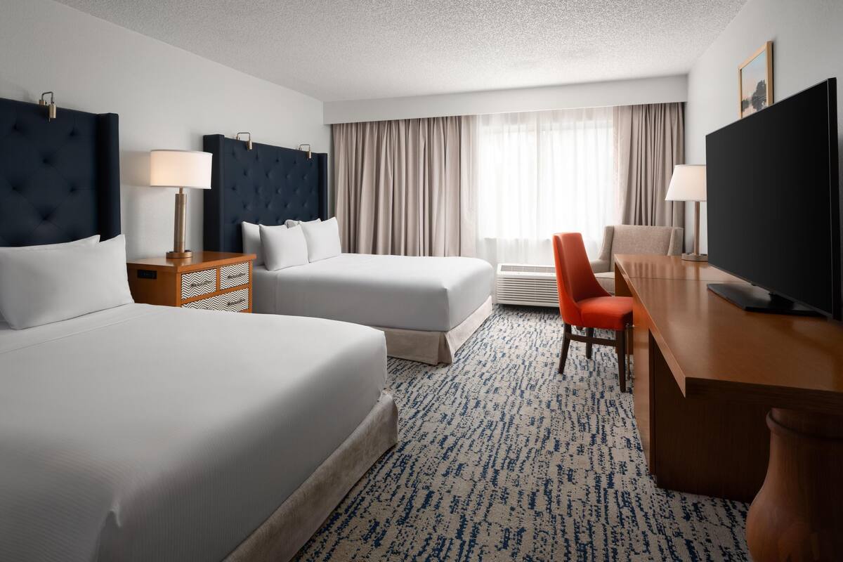 A hotel room with two beds, a desk, a TV, and a window with curtains.