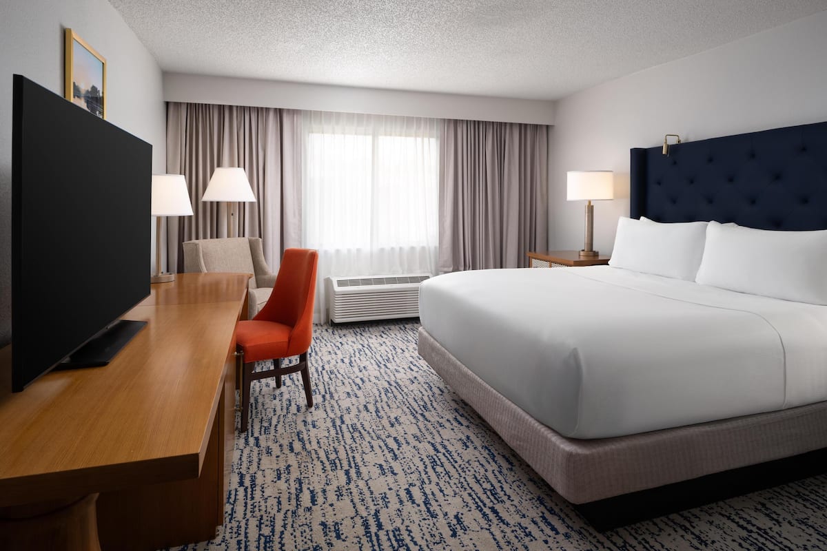 A hotel room with a bed, a desk with a television, a chair, and a nightstand with a lamp.