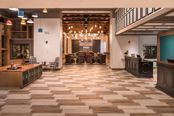 Un vestíbulo espacioso con piso de madera en serpeto, una zona de estar central con una mesa grande y una escalera que conduce a un nivel superior.
