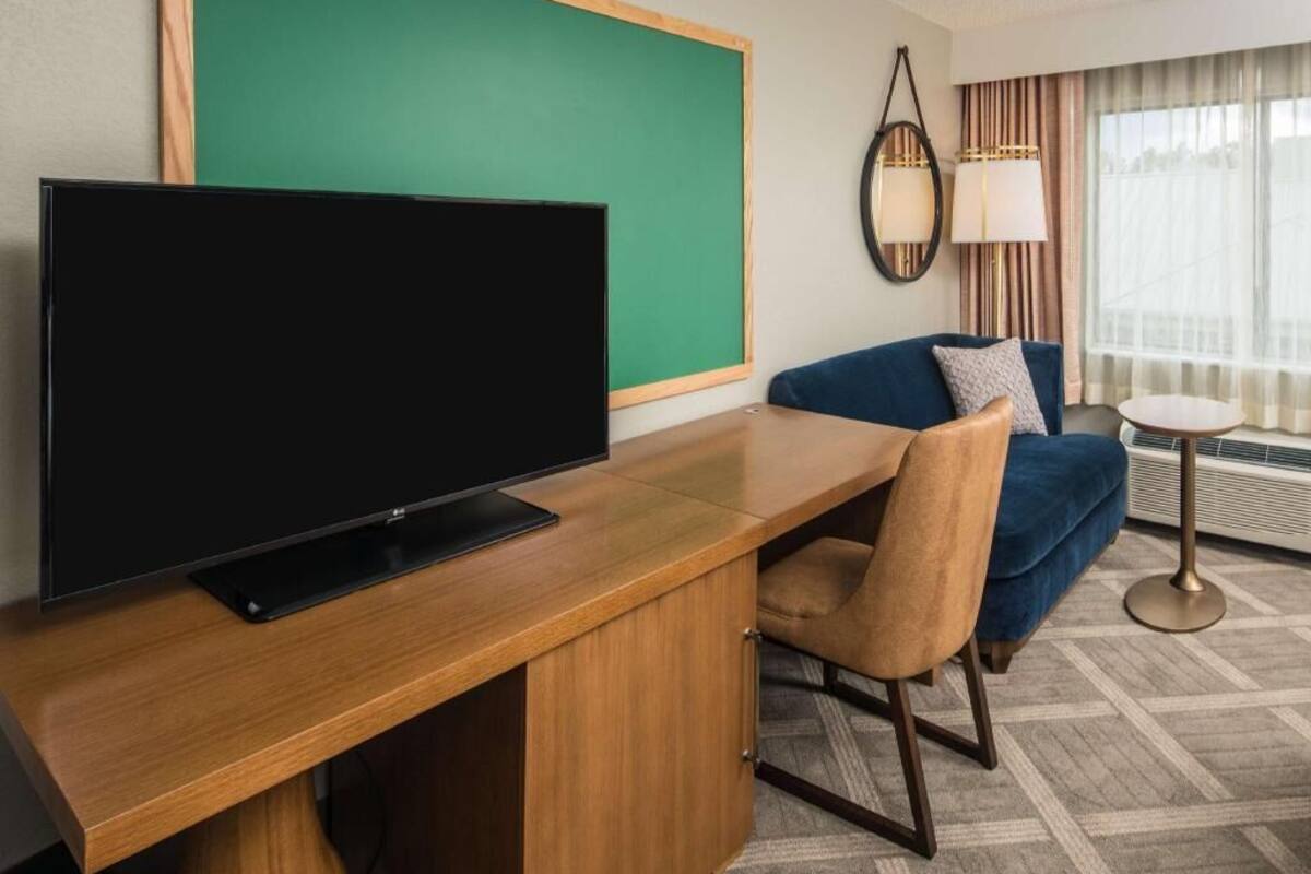 A hotel room with a wooden desk, a television, a blue sofa, a round side table, and a lamp.