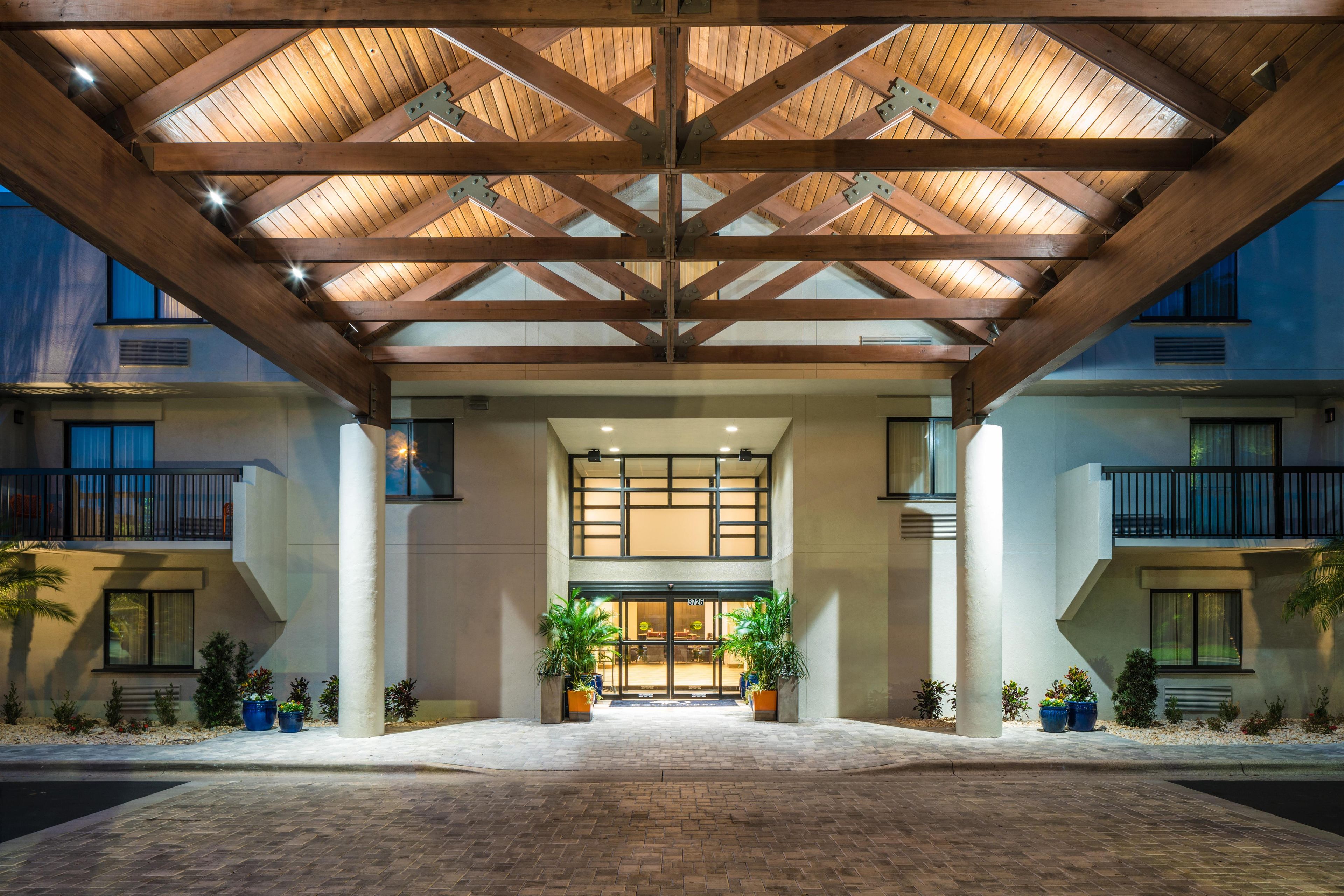 A hotel entrance with a wooden ceiling structure, large windows, and potted plants.