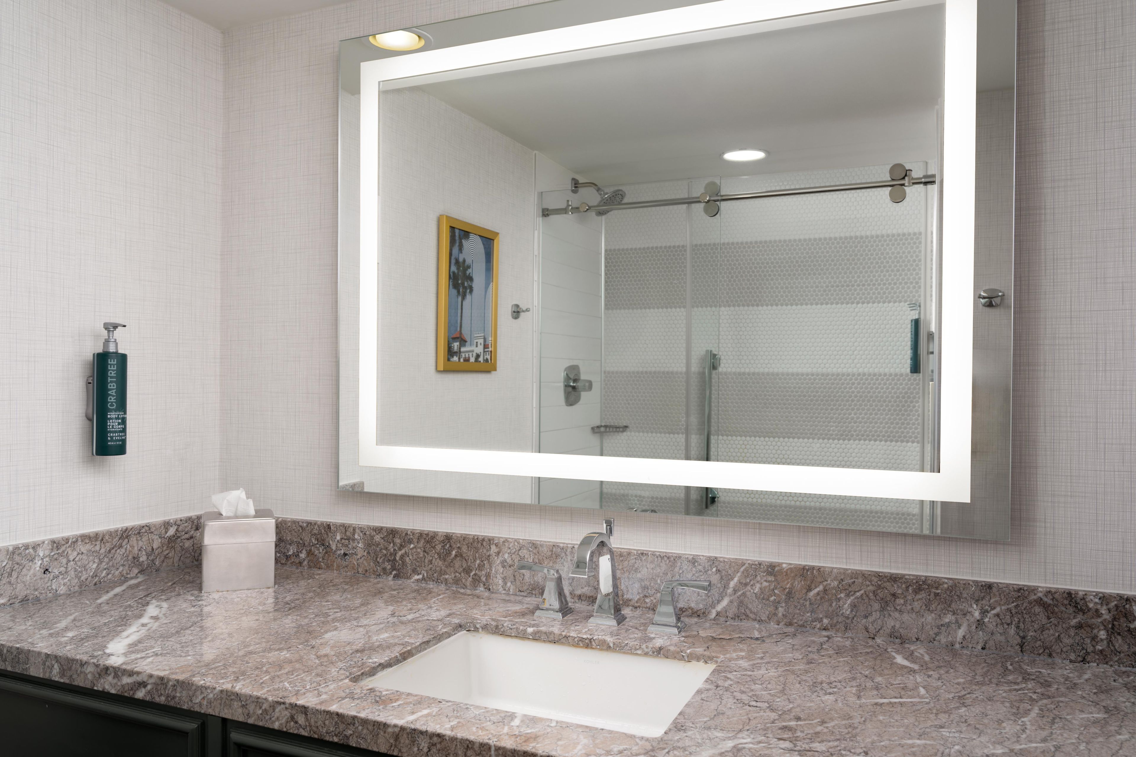 A bathroom with a large mirror, a marble sink, a hand soap dispenser, and a framed picture on the wall.