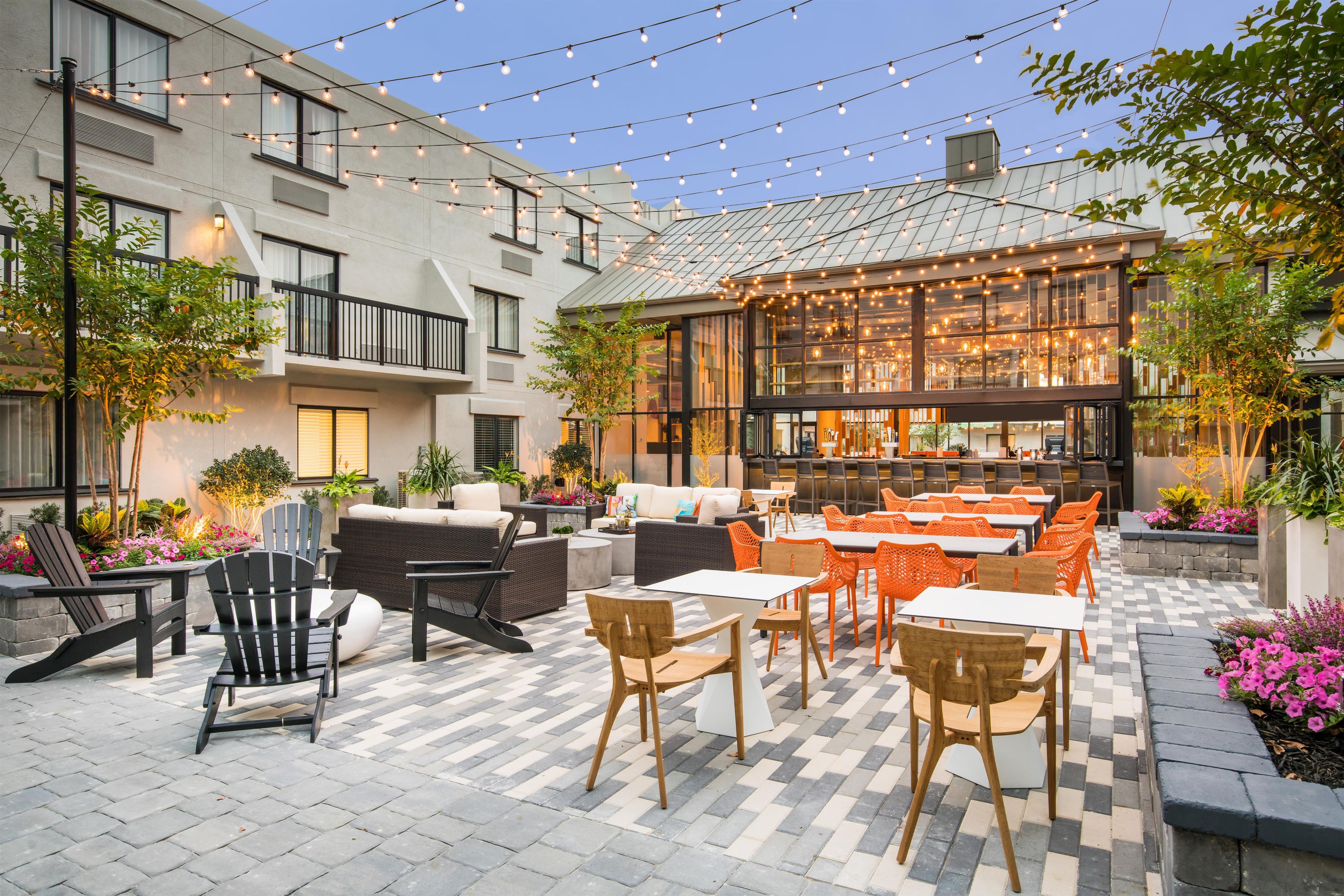 Un patio al aire libre con asientos y mesas, rodeado de edificios y luces colgantes.