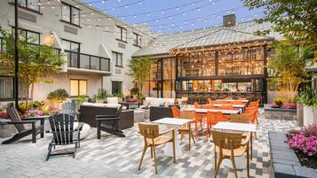 Un patio al aire libre con asientos y mesas, rodeado de edificios y luces colgantes.