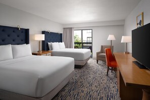 Habitación de hotel con dos camas, un escritorio, una silla, televisión y balcón con cortinas.