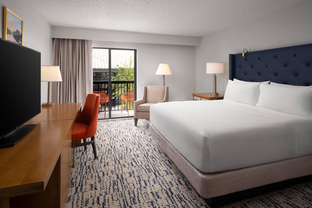 A hotel room with a large bed, a desk with a TV, two chairs, a balcony with a view, and two lamps.