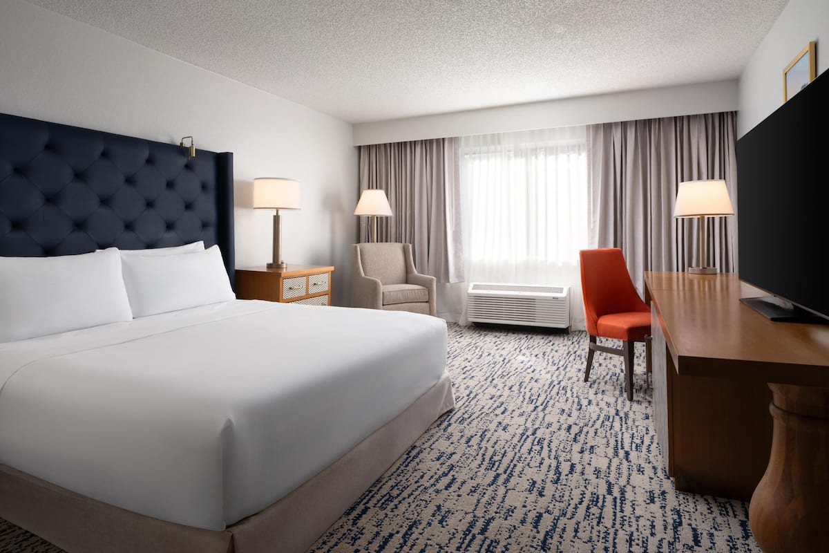 A hotel room with a large bed, a desk, two chairs, a television, and a window with curtains.
