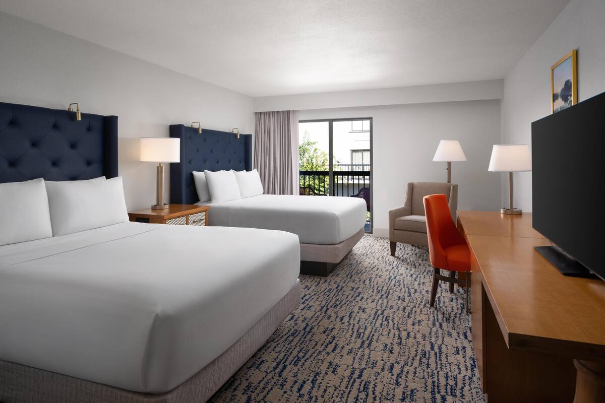 A hotel room with two beds, a desk, a TV, and a balcony with a view.