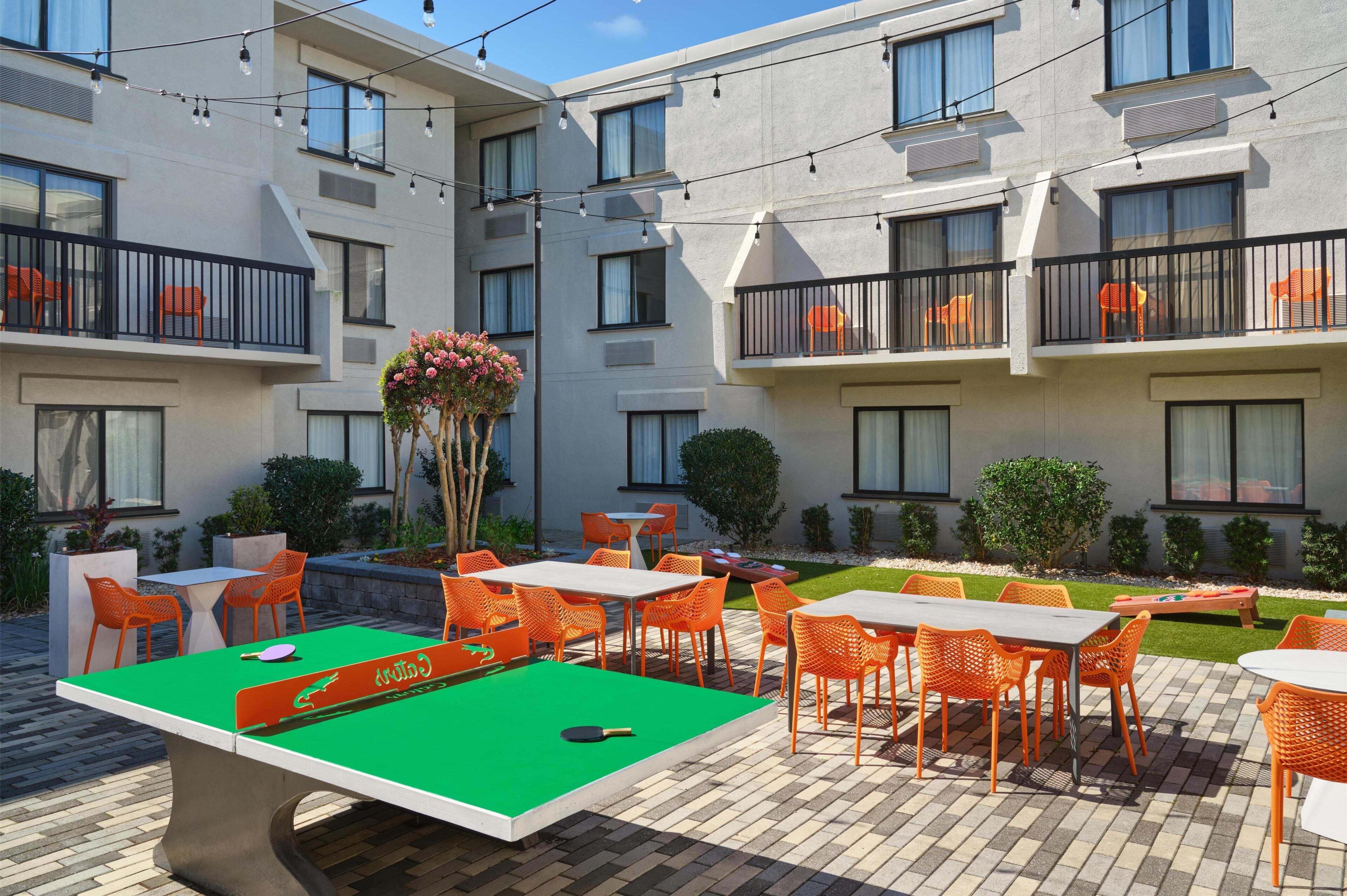 A pool table with orange chairs, a ping pong table, and a small garden area with potted plants and a wooden bench.