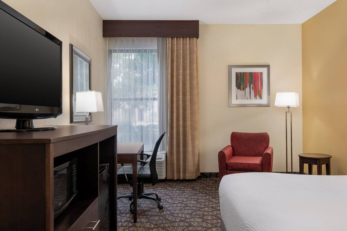 A hotel room with a bed, a desk with a chair, a television, a lamp, and a framed picture on the wall.