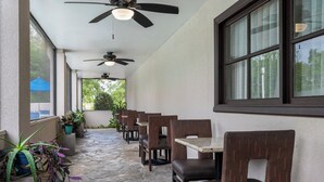 Un espacio de estar al aire libre cubierto, con suelo de piedra, varias mesas y sillas, ventiladores de techo y plantas en macetas.