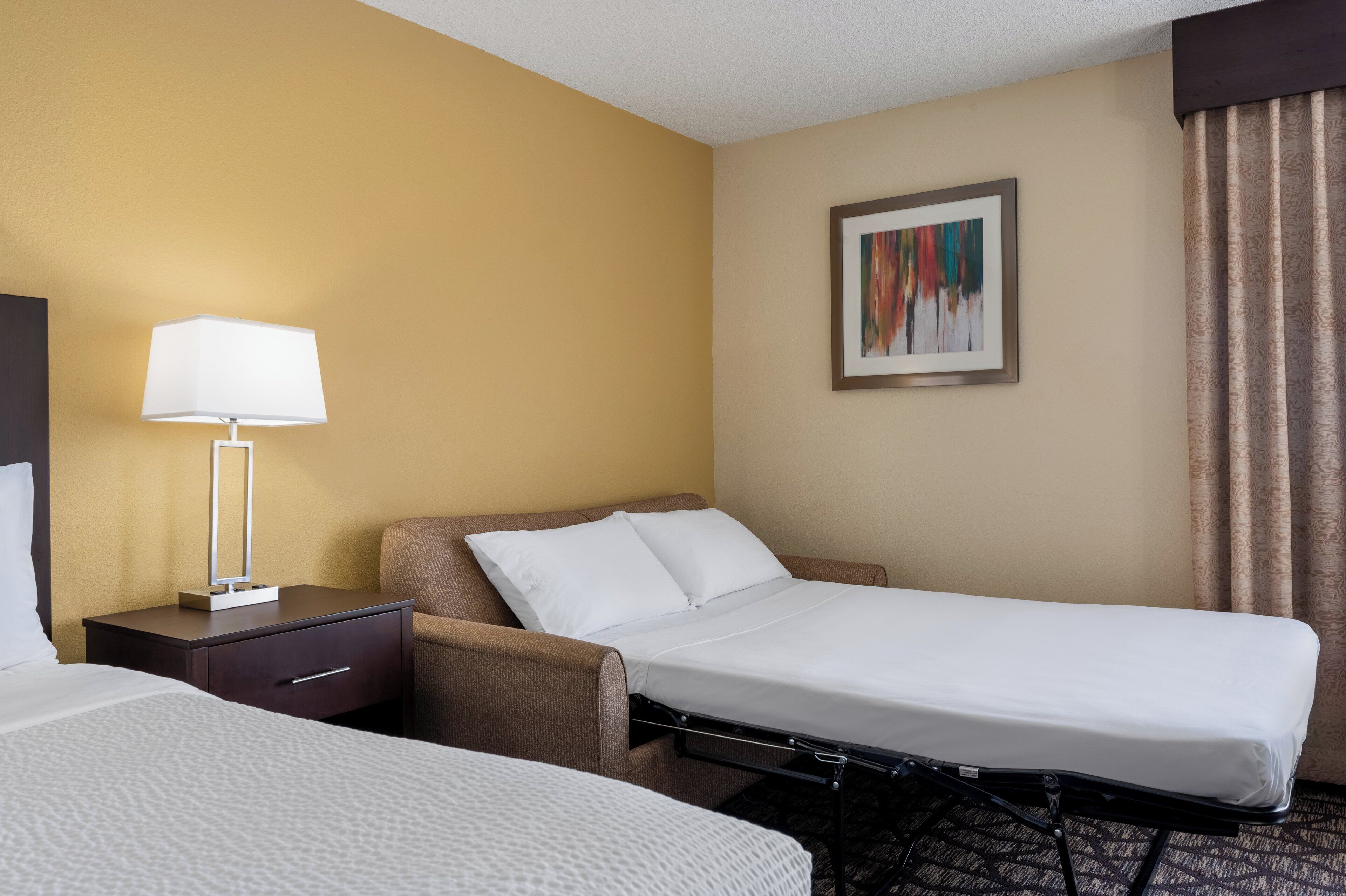 Une chambre d’hôtel avec un lit simple, un petit bureau avec une lampe, une chaise et un tableau au mur.