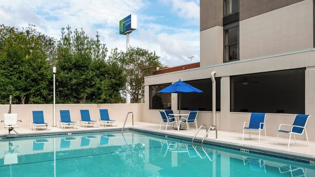 A swimming pool with blue chairs and umbrellas, surrounded by a building with large windows and a sign.