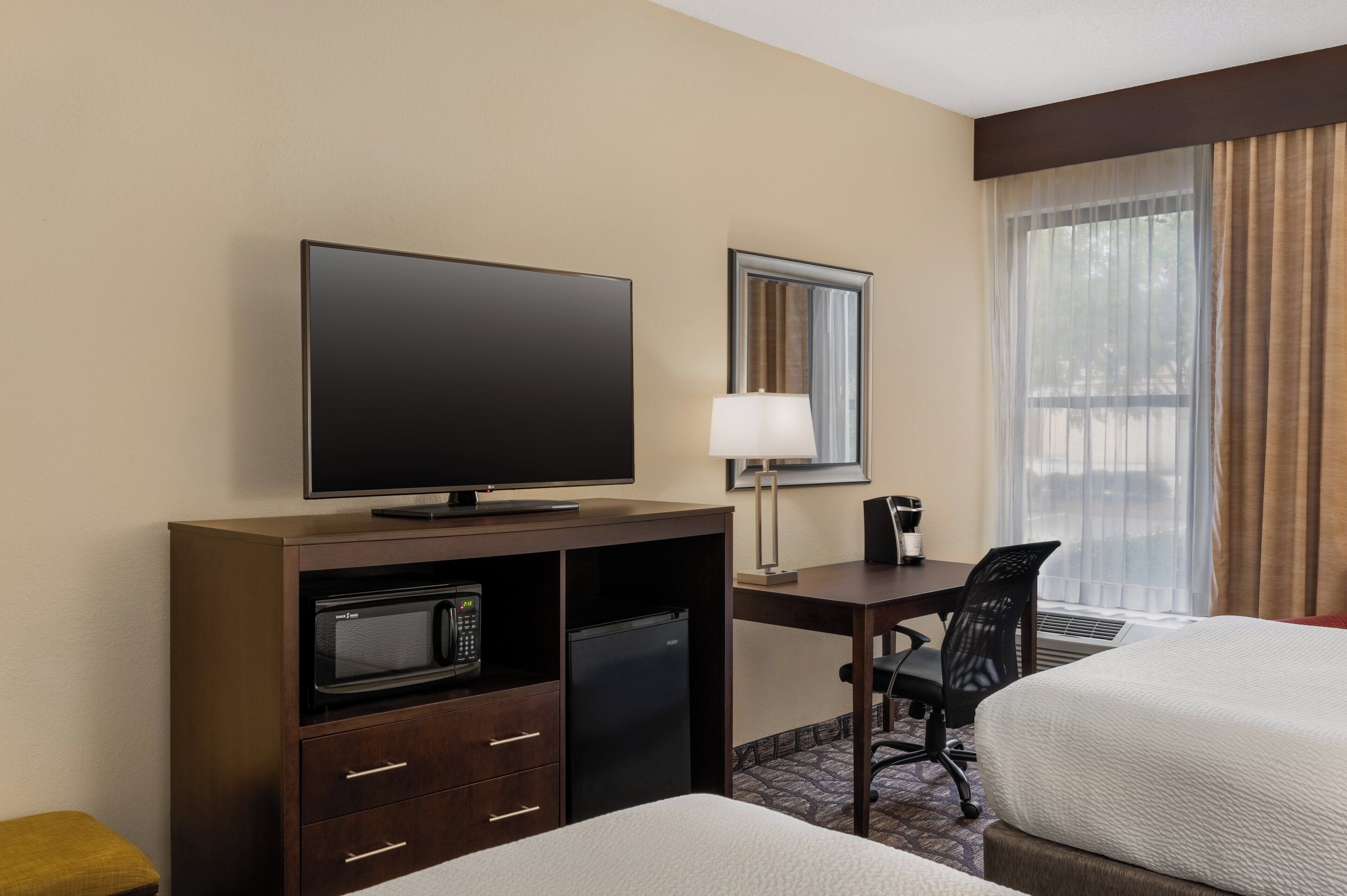 A hotel room with a flat-screen TV, a desk with a lamp, a microwave, a refrigerator, a chair, and two beds.