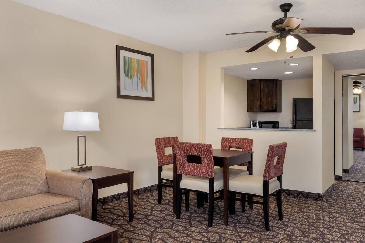 A hotel room with a dining area, a sofa, a lamp, a ceiling fan, and a framed picture on the wall.