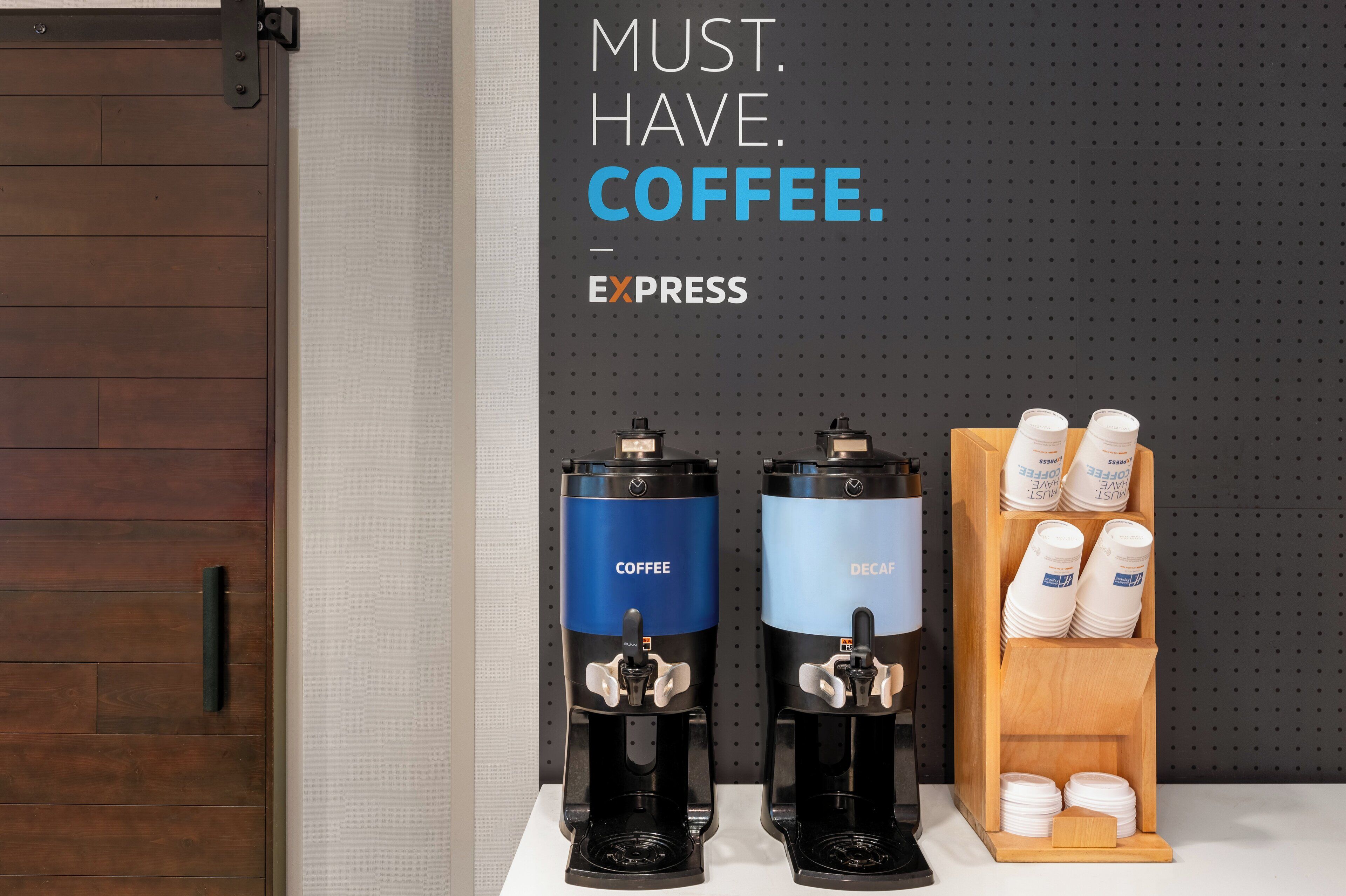 Dos dispensadores de café, uno con la etiqueta "Coffee" y otro con la etiqueta "Decaf", junto a una vitrina de madera con tazas de café.