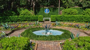 Un jardín con una fuente central, rodeado de parterres florales y una estatua.