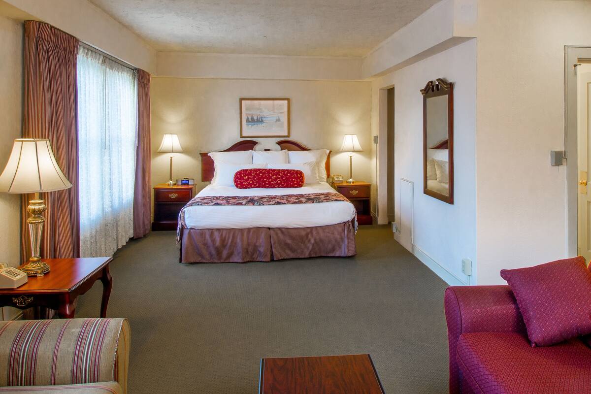 A hotel room with a large bed, two bedside tables with lamps, a red sofa, a striped armchair, and a wooden coffee table.