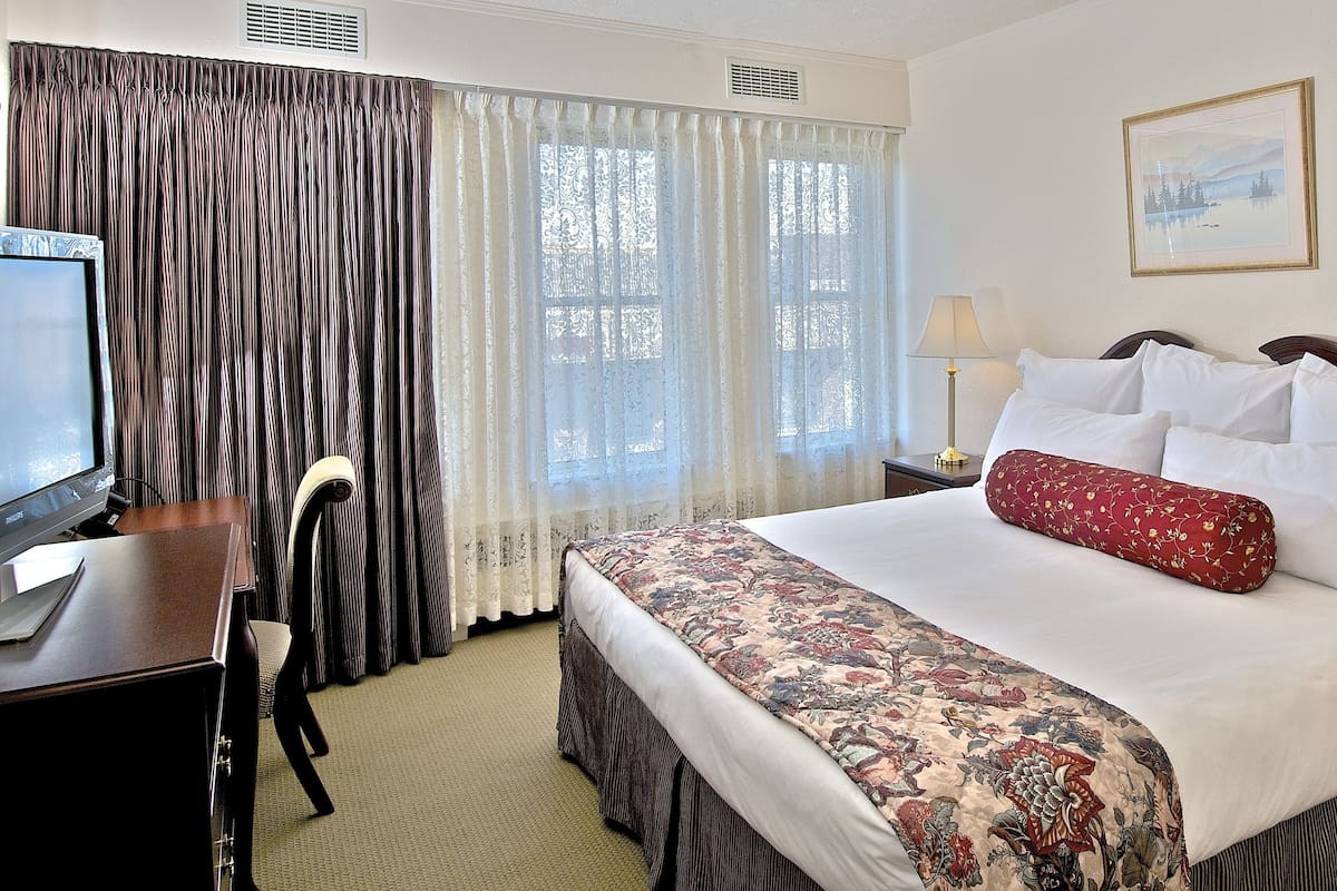 A hotel room with a bed, two bedside lamps, a desk with a computer, a chair, and a window with curtains.