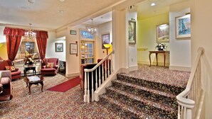Un salon spacieux, doté de meubles recouverts d’un tissu rouge, d’un lustre et d’un escalier recouvert d’une moquette à motifs menant à une autre pièce.