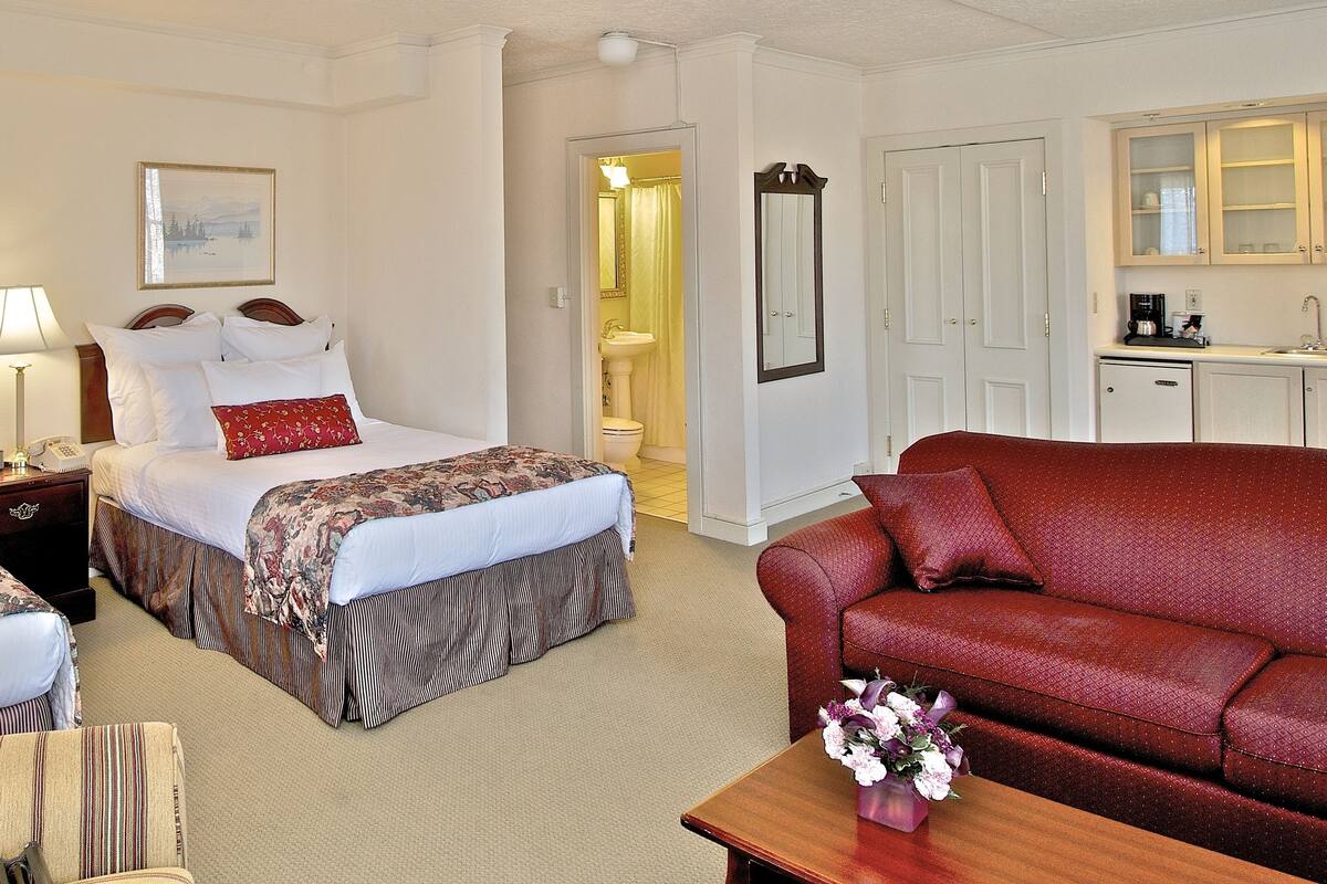 A hotel room with two beds, a sofa, a small table with a vase, and a bathroom visible through an open door.