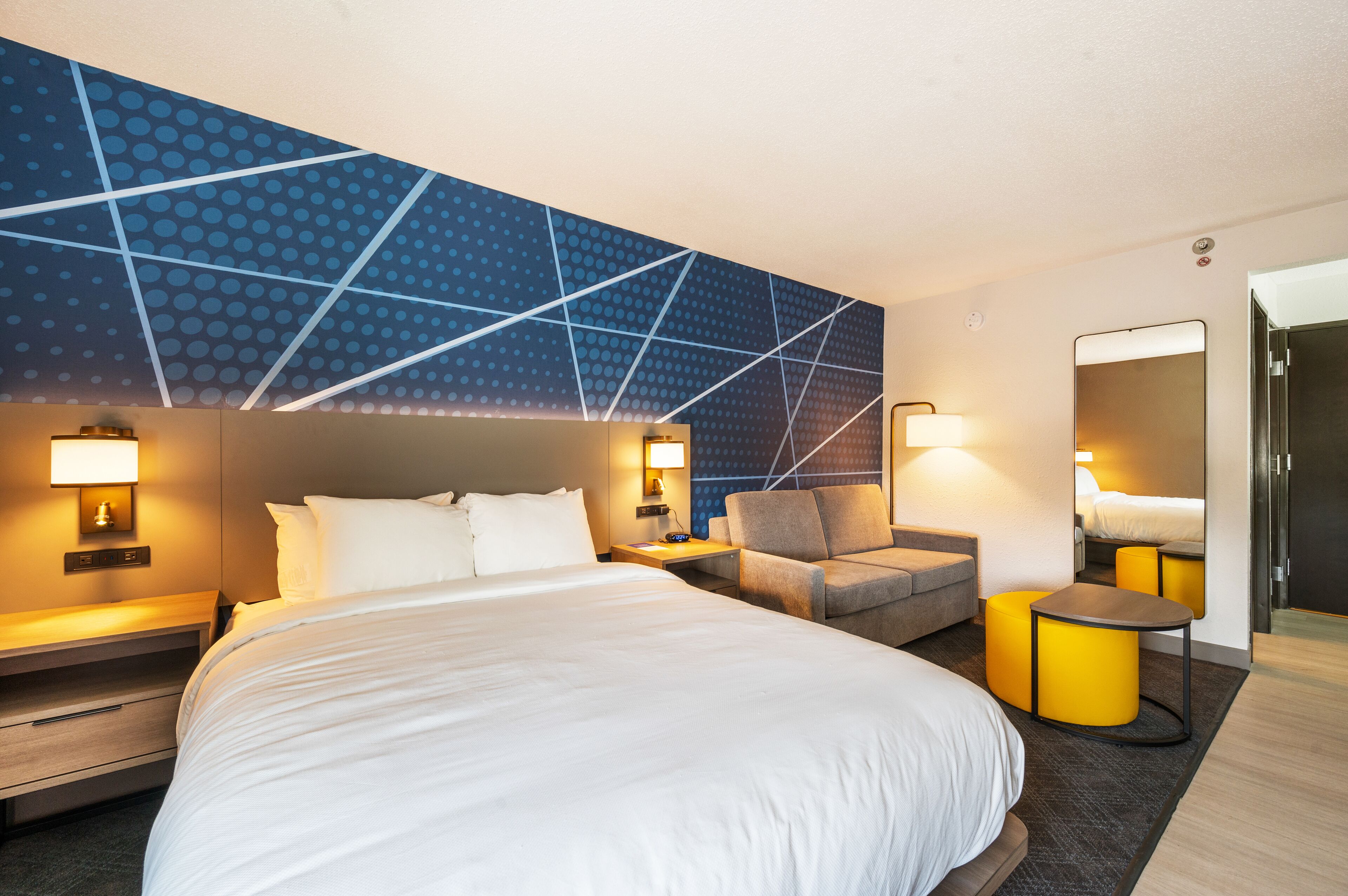 Una habitación de hotel moderna con una cama grande, una zona de estar con una silla y un reposapiés, una mesita y un espejo amplio.